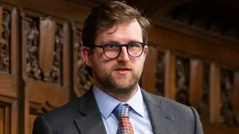 Freddie van Mierlo is standing in the house of commons holding a piece of paper with both hands. He is wearing a light grey suit with a light blue shirt. His tie is multicoloured and he is wearing glasses with black frames. 