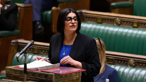 PA Media A woman with glasses, dark hair and a navy suit jacket and blue top, Shabana Mahmood, stands in the House of Commons, a large debating chamber with green leather benches.