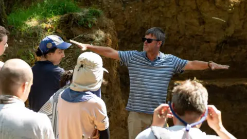 Nick Ashton/British Museum Prof Nick Ashton giving talk at an excavation site. He is talking as he raises his arms out to the side as part of a demonstration. People watch and listen to him. He has short grey hair and wears a blue and white striped top with sunglasses.