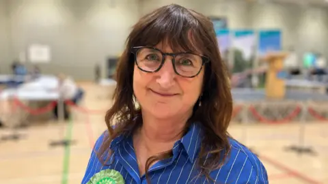 Alex Dunlop/BBC Lucy Galvin, a woman who is looking directly at the camera and smiling. She is wearing a blue and white striped shirt with a green rosette pinned on. 
