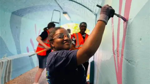 Johana Plazas Sheryl is painting a red streak on the wall with paint clear glasses covering her eyes.
