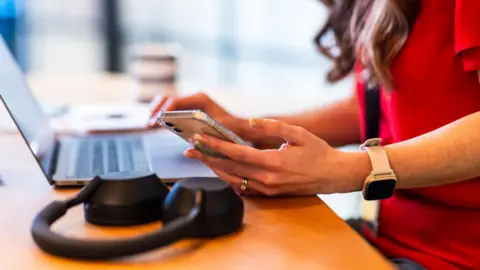 Stock image of a businesswoman holding smart phone near laptop in office.