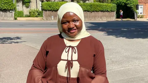 Fadila Issah Fadila Issah standing on a Cambridge residential street on a sunny summer's day. She is wearing a cream hijab and a largely brown long sleeved dress. She is smiling broadly. 