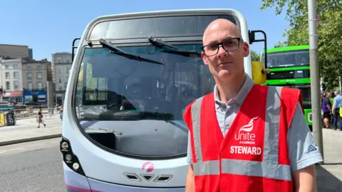 A man in a red tabard over a grey short-sleeved shirt stands next to a First Bus in the centre of Bristol. The bus is mostly grey with some purple and white sections