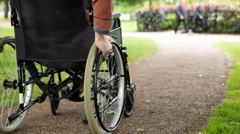 Close-up of a hand on a wheelchair on a path with walkers in the distance