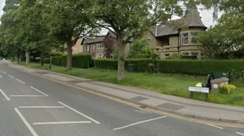 A semi-residential street with wide grass verges and large houses. To the the right a white street sign reads 'Knaresborough Road'.