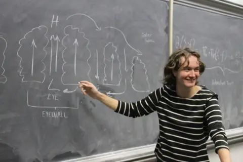 Courtesy of Audrey Palmer Meteorology student Audrey Palmer points to a weather diagram on a blackboard with a piece of chalk while smiling and facing a classroom. 
