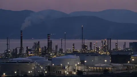 A view of the Motor Oil refinery in Agioi Theodoroi near Corinth, Greece during the evening. Behind the refinery, the sea and hills are visible but dark.