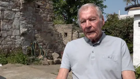 A man in a grey collared shirt in a garden with a stone wall behind him.