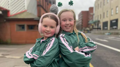 Two girls are standing with their arms folded and their backs resting against each other. They're smiling and both are wearing matching green jackets. One has ear muffs on, the other has a shamrock hairband on. 