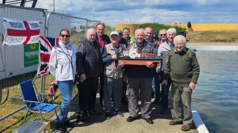 About a dozen members of Redcar Model Boat Club smiling next to the partially refilled lake. A member in the centre of the group is holding a brown and white model boat. There's a flag on the side of a portable building next to them, with the name of the club on it.