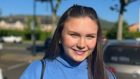 Maria is smiling at the camera. She has long brown hair and is wearing a bright blue hoodie.