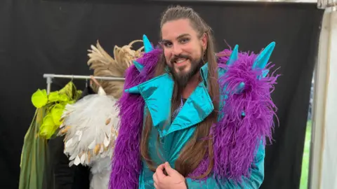 Adam stands inside a tent wearing a vibrant costume that includes a shiny turquoise jacket with large lapels and blue spikes on the shoulders, with a purple shawl. Behind them is a clothing rack with other colourful costumes, including one with white feathers and another with bright green accents. The background is dark, highlighting the vivid colours of the outfits.