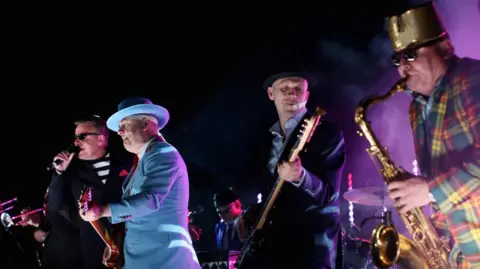 PA Media Four men in a band performing in darkness. They are wearing multicoloured outfits.