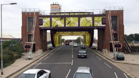 A view of Drypool Bridge, which has two large brick towers, and metalwork covered in Venn diagram inspired designs. Cars drive across the structure, which also features two red and white striped safety barriers.