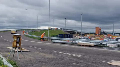 Northumberland County Council A large modern road bridge sweeps over a new railway station with construction workers wearing orange hi-vis jackets and trousers working on the barriers. Under the bridge construction work with diggers continues 