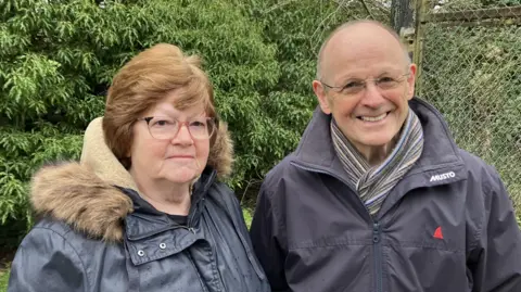 The image shows two people standing in front of a lush green bush. On the left is a woman wearing a black coat with a fur-trimmed hood and glasses. On the right is a man dressed in a black coat, a striped scarf, and glasses.