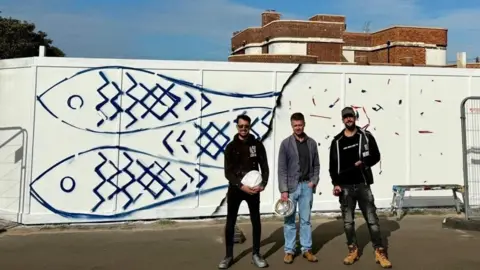 Debby Besford Three men stand by the hoarding outside the Iron Duke pub. The hoarding is painted white with three herring painted in blue depicted.
