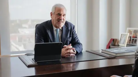 Podhurst Orseck A man with grey hair, wearing a navy suit, sat at a wooden desk against a window with an ipad 