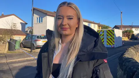 Carla Fowler/BBC A woman with blonde hair and a black coat stands on a street which is cordoned off after a gas explosion 