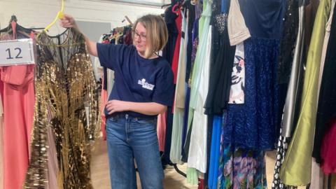 A NEST volunteer holding up a gold and black dress in front of two racks full of colourful evening dresses 