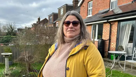 Liz Lockwood wears a yellow coat and a pale pink jumper. She is looking at the camera, wearing sunglasses and is standing in a garden, with homes visible behind her as well as a bird bath and a garden table and chair