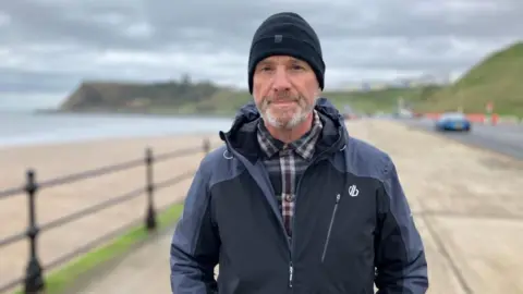 BBC/Seb Cheer A man, Paul, looks into the camera with a neutral expression on his face. He is wearing a coat and beanie hat. Scarborough Castle is in the background.