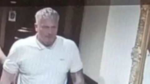 A standing man with blonde hair. He is wearing a white polo shirt and has a tattoo sleeve on his left arm.