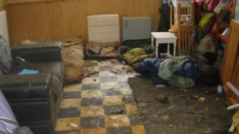 RSPCA Room with dirty looking dog beds, sofa and rubbish strewn across the floor with chairs stacked up in one corner