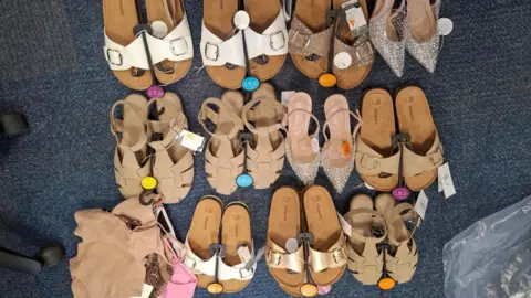 Policing Peterborough A large row of summer shoes and sandals, that have labels on them, and hangers. A pile of clothes is to the bottom left. All the items are on grey carpet. Chair legs are to the left and a plastic bag to the right. 