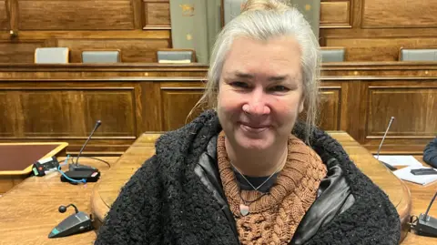 Woman smiling. She has grey hair in a bun. She's wearing a brown jumper and a fluffy back jacket over the top. Behind her, there's a brown wooden table with microphones on it. In the back, you can see a wooden panel with 5 chairs behind it. 