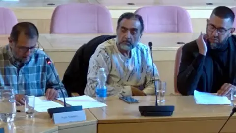 Peterborough City Council Ali wearing a blue checked shirt looking at a paper- Nawab wearing a white t-shirt with black lines and Zameer Ali in a black suit sitting in the meeting 