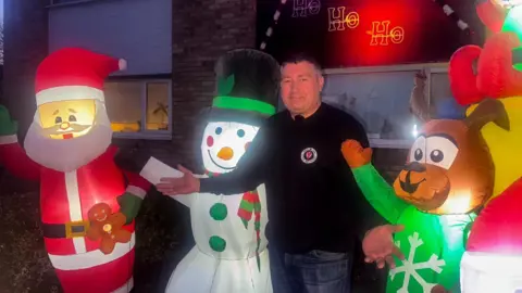 BBC/ Emily Johnson A smiling man stands outside a house decorated for Christmas, surrounded by large inflatable figures: Santa holding a gingerbread man, a snowman in a green hat and scarf, and a reindeer in a green snowflake jumper. The house behind them is lit with festive lights and a glowing “Ho Ho Ho” sign. The man shrugs with his arms up.