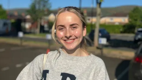 Ella-Beth is smiling at the camera. She has blond hair ties back. She is wearing  grey jumper.