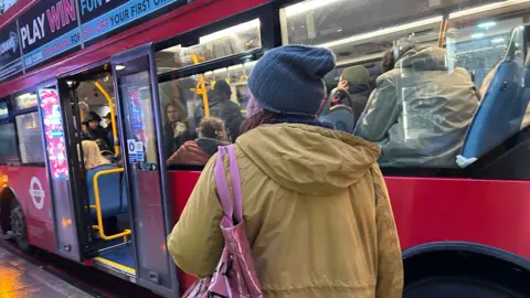 BBC A woman in a coat stands facing away from the camera next to a London bus