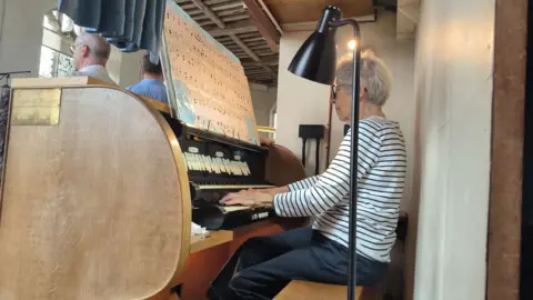 Cressi Sowerbutts An elderly woman playing the organ in a church. She has short grey hair, glasses and is wearing a white and black striped top with navy blue trousers. She is reading enlarged sheet music as she performs. 