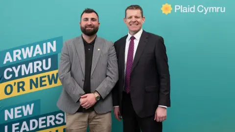 Plaid Cymru Oli Bradley-Hughes standing alongside Plaid Cymru leader Rhun ap Iorwerth in front of party branding including the Plaid Cymru logo, a bright yellow Welsh poppy. The words "new leadership for Wales" are partly obscured behind Bradley-Hughes. Both men are looking at the camera and smiling. 