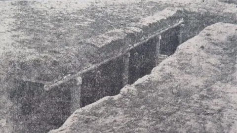 The Bedfordshire Standard A black and white picture of a World War One trench which has wooden posts on one side. It is a grainy image.