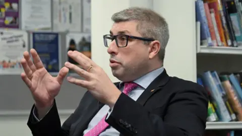 Reuters Mark Arnull in a dark suit, light blue shirt and pink tie. He is looking to the side, while raising his hands in the air. He sat in a library in front of a book shelf.