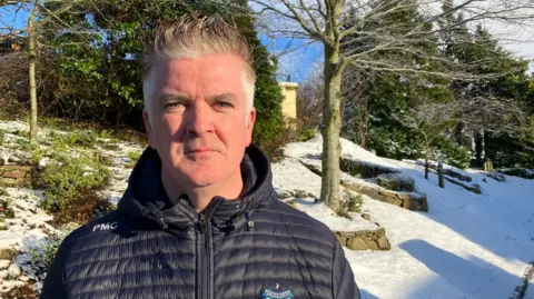 Headmaster Philip McCullagh stands in front of his school's grounds which are covered in snow. He has grey hair and is wearing a black coast