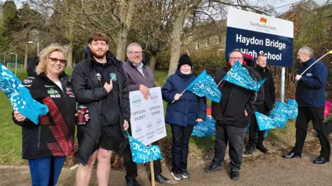 Education workers and supporters holding National Education Union flags and an official strike picket sign outside Haydon Bridge High School. There are seven strikers standing next to a sign at the entrance to Hayden Bridge High School.