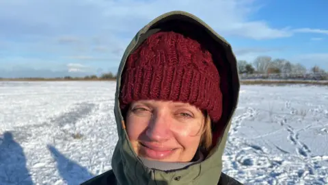 John Devine/BBC Laura Upton is smiling at the camera. She is standing on a snowy and icy field and is wearing a green coat with the hod up over a red woolly hat.