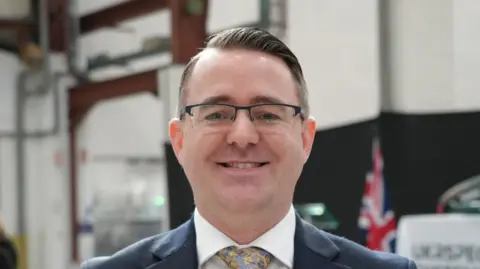 Shaun Whitmore/BBC A head and shoulders image of a man wearing glasses and a navy blazer over a white shirt with a tie. He is smiling and looking directly into the camera.