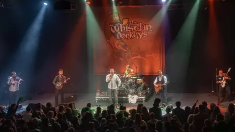 The band playing a range of instruments on stage with a red poster with 'The Whilstin Donkeys' written on it.