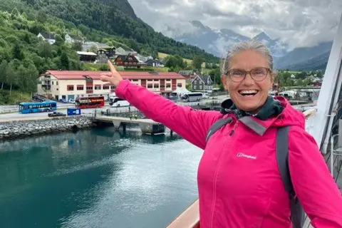 Scott Hastings Jenny Hastings, wearing a pink jacket and standing on a boat in the foreground, smiles and points to houses nestled in a valley