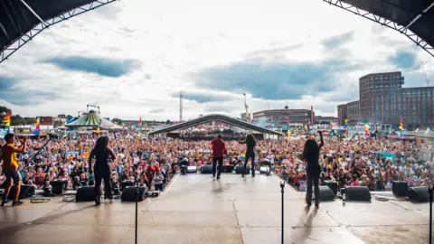 Birmingham Pride Thousands of people attending Birmingham Pride at Smithfield, as presenters introduce acts to the main stage. A sea of people leads off from a stage with tower blocks in the distance.
