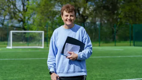 Julie Norris with short brown hair, blue jumper and black clipboard smiling and while standing on a football pitch. There is a goalpost and some trees in the background.