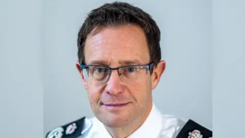 Staffordshire Police A man with short black hair, glasses and wearing a police uniform, smiles while standing in front of a light background.