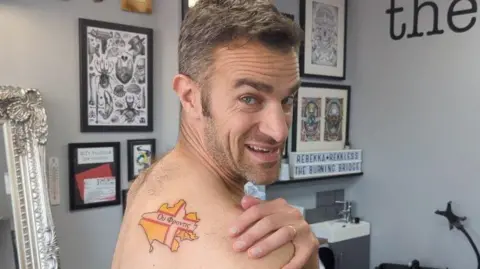 Steve Harris showing off a tattoo on the back of his shoulder. The tattoo is in the shape of the map of Dorset and is coloured yellow, with a white and red cross, which is the Dorset flag.