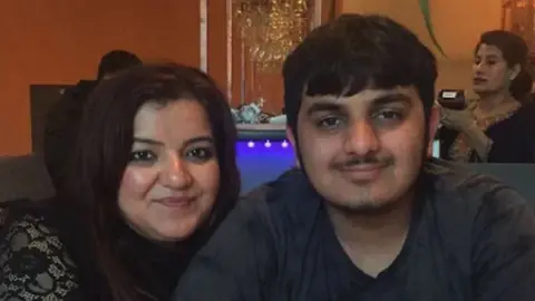 Family Pooja and Ronan sit at a table in a restaurant. He is wearaing a grey t-shirt. A woman can be seen behind them holding the card machine.